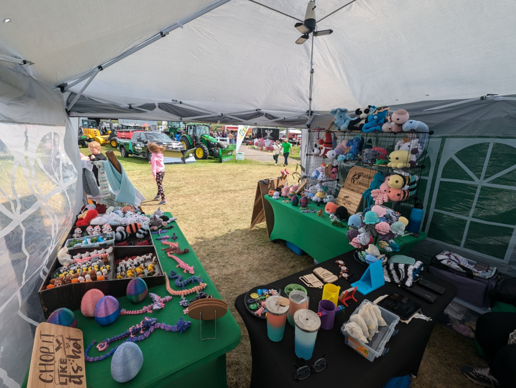 A vendor stall at the Orono Fair, set up under a white canopy tent, featuring tables covered in green and black cloths filled with handmade crocheted plushies, 3D-printed articulated lizards, and various craft items, with an outdoor fairground scene visible in the background.